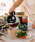 Person pouring coffee from borosilicate glass carafe of 12 Cup Drip Coffee Maker With Auto Off And Reusable Filter into mug in kitchen