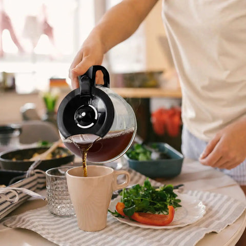 Person pouring coffee from borosilicate glass carafe of 12 Cup Drip Coffee Maker With Auto Off And Reusable Filter into mug in kitchen