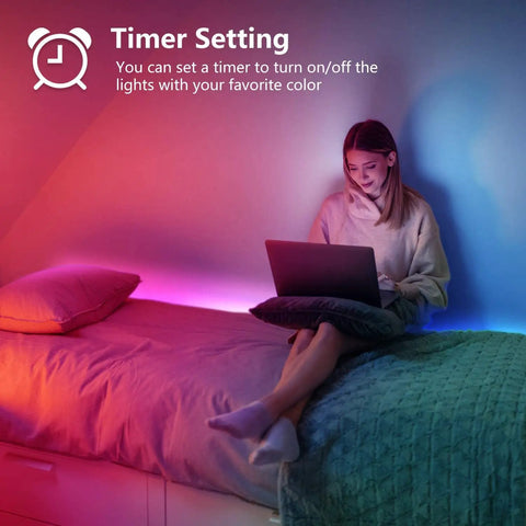 Woman using a laptop in a bedroom with 65 Foot Color Changing LED Light Strip With Remote And App Control For Bedroom Party Setup Dope RGB Lighting With 24V Power Supply Indoor Mood Lighting For Home Decor And Music Sync installed on the wall.