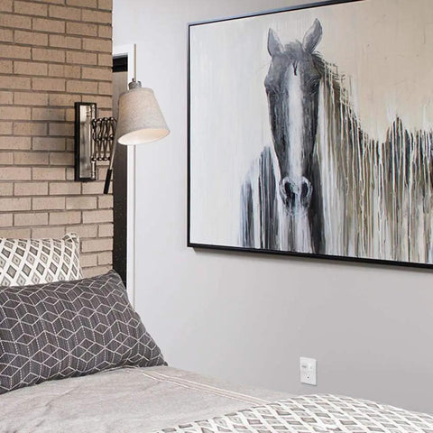 Bedroom with abstract horse painting on wall, grey geometric bedding, white lamp and brick wall detail.
