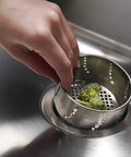 Hand placing broccoli in stainless steel 3-in-1 kitchen drain flange with removable lid, water-holding cap, and sink strainer.