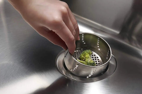 Hand placing broccoli in stainless steel 3-in-1 kitchen drain flange with removable lid, water-holding cap, and sink strainer.