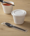 Six Point Five Inch Wrapped Compostable Spoons For Hot And Cold Food 250 Individually Wrapped Plant Based Black CPLA Disposable Utensils BPI Certified Eco Friendly Cutlery For Restaurants And Events, wrapped black spoon beside soup cups