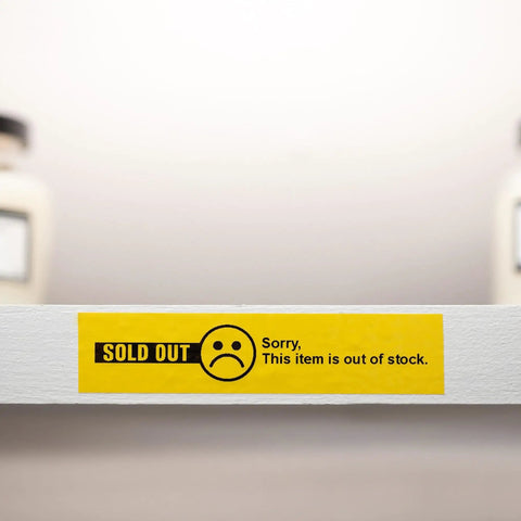 Yellow 'Sold Out' label with sad face icon and text 'Sorry, This item is out of stock' on a white shelf with blurred background.
