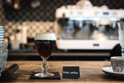 Glass of Irish coffee on a table with a mini chalkboard sign labeled 'Irish coffee', blurred coffee machine in the background.