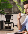 Heavy Duty Fully Automatic Espresso And Drip Coffee Maker With Digital Display Six Steam Nozzles For Killer Brews 10 Cup Glass Carafe in use with a man drinking coffee.