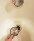Hand holding a stainless steel Bathroom Sink And Tub Hair Catcher 2 Pack Non Slip Drain Protector Anti Clog Filter above a tub drain.