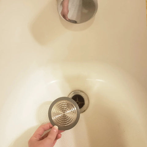 Hand holding a stainless steel Bathroom Sink And Tub Hair Catcher 2 Pack Non Slip Drain Protector Anti Clog Filter above a tub drain.
