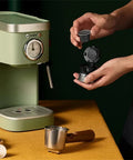 Hand holding a coffee capsule near a Heavy Duty No Hassle Electric Cappuccino Maker With Smooth Pour Milk Frother.