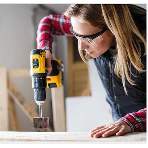 Front view of a woman in safety glasses using a yellow cordless drill with a flap wheel sanding attachment on wood.