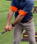 Golfer demonstrating Orange Golf Swing Trainer Aid For One Piece Takeaway And In Sync Swing Keeps Arms And Body Connected on green field.