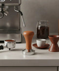 Top Shelf Espresso Coffee Tamper With Polished Stainless Steel Base And Sturdy Wooden Grip on a countertop next to espresso tools.