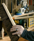 Person holding a large wooden push broom in a workshop setting. Various tools and equipment visible in the background.