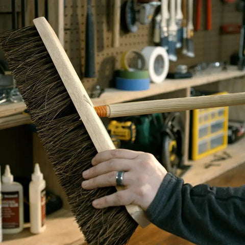 Person holding a large wooden push broom in a workshop setting. Various tools and equipment visible in the background.