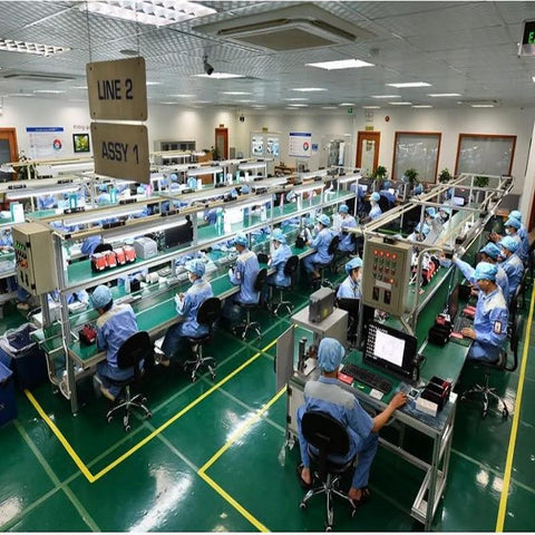 Wide view of electronics assembly line; workers in blue cleanroom outfits at long benches.