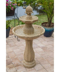 Stone outdoor fountain with tiered design on a patio, surrounded by plants and a pond in the background.