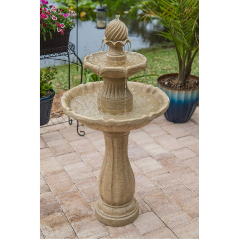 Stone outdoor fountain with tiered design on a patio, surrounded by plants and a pond in the background.
