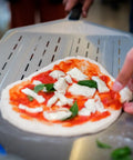 Person using a 14 Inch Perforated Pizza Peel With Long Handle Aluminum Lightweight Pizza Paddle for turning pizza on stone ovens.
