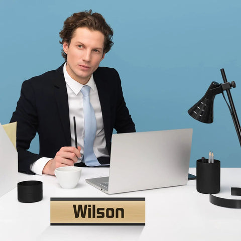 Professional desk setup with a man at a laptop beside a 2x8 inch name plate holder displaying 'Wilson', part of 12 Pack Aluminum Desk Name Plate Holders 2 By 8 Inch Blank Name Plates For Office School Classroom Black Durable Metal Display Stands To Show Off Your Title And Keep Desk Neat At End Of Year, black finish.
