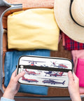Hands holding Go To Travel Toiletry Bag For Women And Men Foldable Dopp Kit with aircraft pattern next to packed suitcase
