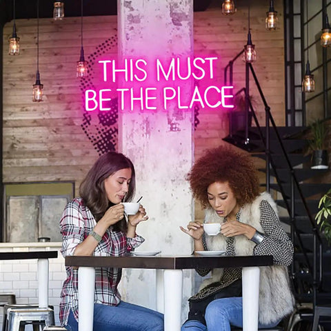 Two friends in a cafe with 'This Must Be The Place Neon Sign Pink Wall Decor For Bedroom And Party Vibes' in background.
