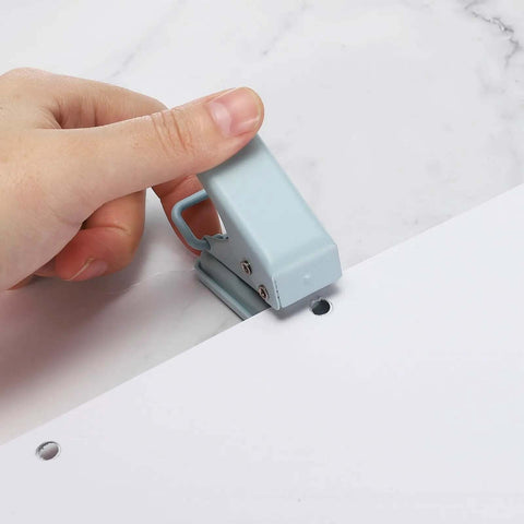 Hand using blue handheld hole punch on paper, representing Pink Handheld Heavy Duty Hole Punch Set Two Piece Metal Paper Punch For Tags And Tickets DIY Craft Scrapbooking Easy Grip Tool With 6mm Hole Perfect For Card Making School Projects Office Use.
