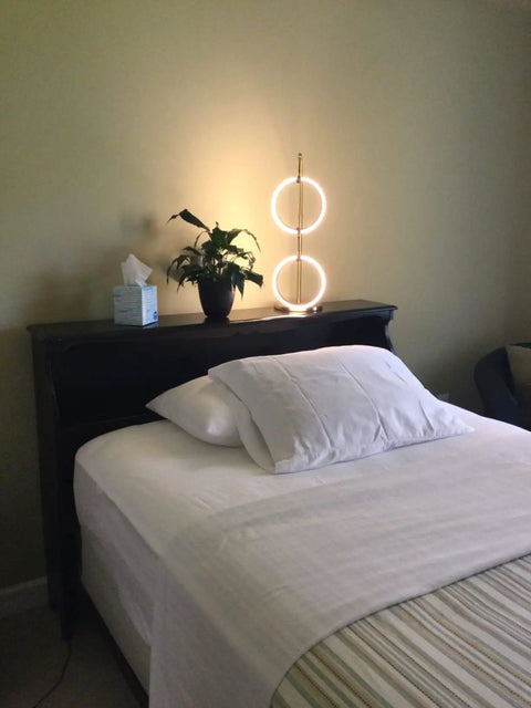 Nova Modern Arc Table Lamp with Two Circle Ring Style Pendant on nightstand beside bed in bedroom setting, brass finish.