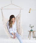 Woman sitting in a macrame boho hammock chair with rope swing, 2 cushions, for indoor and outdoor living, in a modern room.