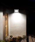 JUDYSSY Solar Flood Lights Led Floodlight 100W installed by a wooden door, providing bright illumination on a wall.