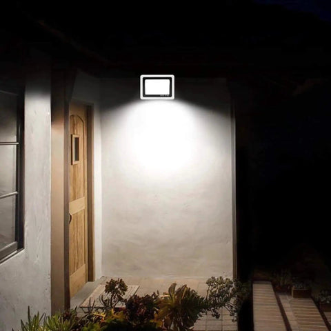 JUDYSSY Solar Flood Lights Led Floodlight 100W installed by a wooden door, providing bright illumination on a wall.