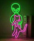 Green alien holding pink cat neon sign glowing on acrylic base with books in background