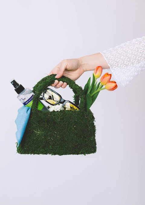 Front view of a moss-covered handbag with a Flents Wipe N Clear spray bottle, sunglasses, blue cloth, and orange tulips.