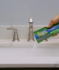 Hand holding Drain Opening Pacs For Clogged Toilets Sinks Showers And Tubs Hair Clog Remover above bathroom sink.