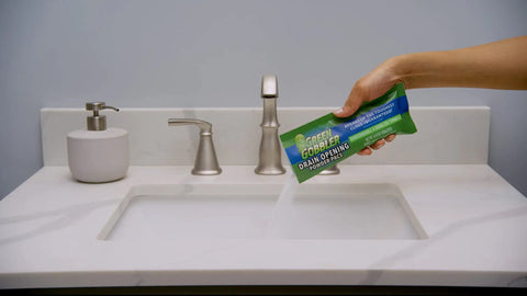 Hand holding Drain Opening Pacs For Clogged Toilets Sinks Showers And Tubs Hair Clog Remover above bathroom sink.