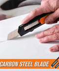 Side view of retractable utility knife with orange grip and carbon steel blade cutting cardboard.