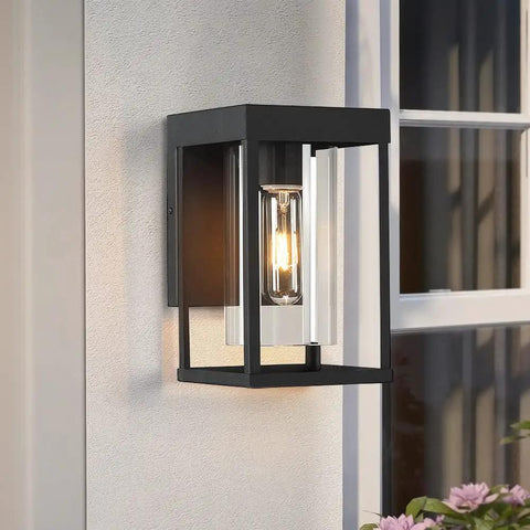 Single 9-inch tall black outdoor wall sconce with clear glass shade mounted beside a window, illuminating the exterior. 2-Pack Black Outdoor Wall Light Fixtures Exterior Wall Lantern Sconce For Porch And Patio, 9-Inch Tall Wall Mount With Clear Glass Shade, Weatherproof Exterior Lighting For Curb Appeal.