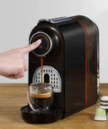 Compact Capsule Coffee Machine For Home Office Auto Espresso Maker One Touch Brew Mini Size Milk Frother Included For Soy Milk And Tea Options Quiet Desk Friendly Coffee Gadget No Fuss, shown with a hand pressing the one-touch button on a wooden surface, capsules beside.