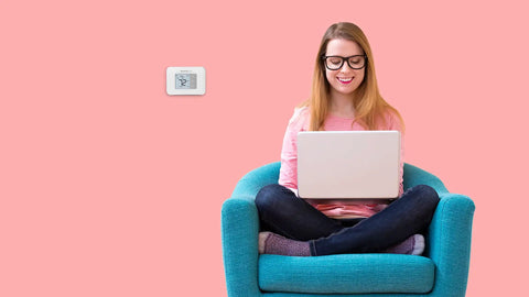 Woman sitting on teal chair near non programmable thermostat with backlit display and no fuss simple manual controls on pink wall.