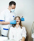 Dentist examining a patient's teeth in a dental office setting, both wearing white clothes and blue gloves, dental equipment visible.