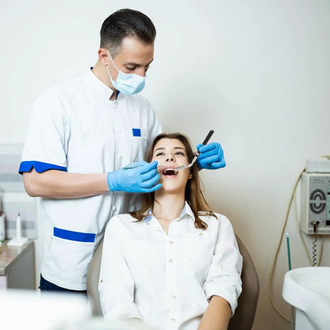 Dentist examining a patient's teeth in a dental office setting, both wearing white clothes and blue gloves, dental equipment visible.