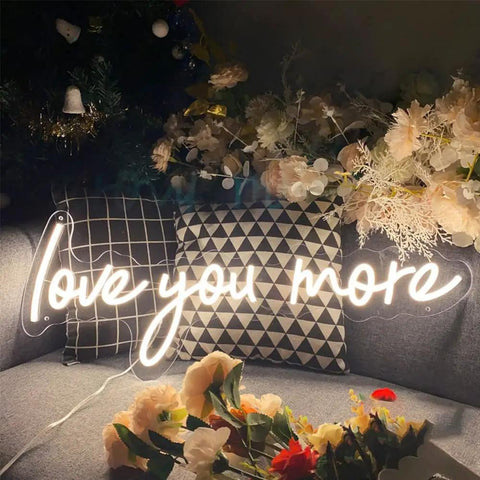Neon Sign In Transparent Acrylic, Dimmable LED Wall Light That Turns Any Room Into A Cozy Vibe, Indoor Decor For Bedroom Living Room Office Or Bar, Perfect For Christmas Parties And Weddings, Warm White 23.6x9 Inches, 'Love You More' glowing among pillows and flowers