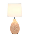 Table lamp with textured peach ceramic oval base and white shade, stands on a reflective surface against a white background.