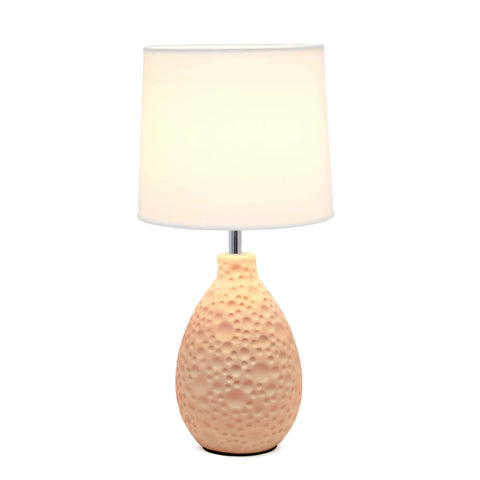 Table lamp with textured peach ceramic oval base and white shade, stands on a reflective surface against a white background.