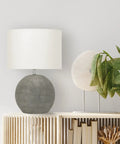 Round Terracotta Table Lamp With Canvas Shade In Distressed Grey Finish, placed on a wooden shelf next to green leaves.