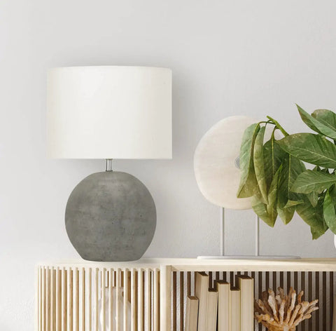 Round Terracotta Table Lamp With Canvas Shade In Distressed Grey Finish, placed on a wooden shelf next to green leaves.