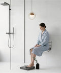Person sitting on 16 Inch Foldable Shower Seat Wall Mounted For Adults With 400 Lb Max Load Easy Drop Down Design Heavy Duty Waterproof Wood And Stainless Steel Build No Fuss Fits Small Inside Shower Spaces White in a modern bathroom.
