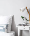 White bedside table with book, glasses, plant, and lamp beside bed with pillow.