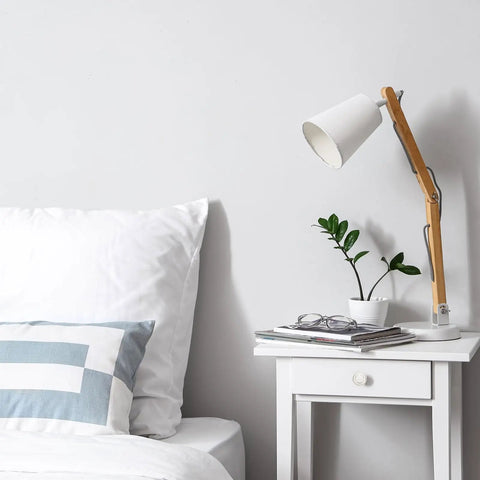 White bedside table with book, glasses, plant, and lamp beside bed with pillow.