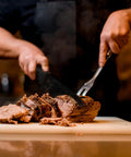 Person slicing cooked meat on a cutting board with a meat fork and knife indoors.