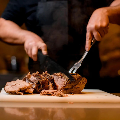 Person slicing cooked meat on a cutting board with a meat fork and knife indoors.
